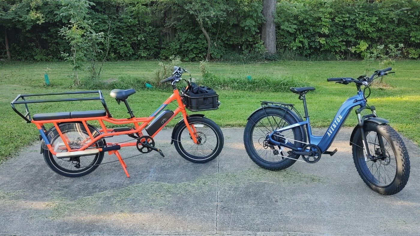 Two e-bikes parked on concrete. On the left is an orange RadWagon 4 from Rad Power Bikes. On the right is a blue Aventure.2 from Aventon.